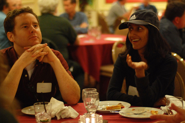 一个女人（在右边）和一个男人（在左边）坐在宴会厅的桌子旁。他们都穿着黑衬衫。女人在说话，脸上带着微笑，一只手向前指，而男人则望向远方，脸上流露出深思熟虑的神情。