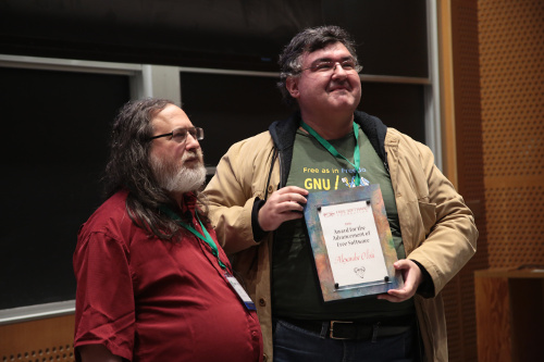 Richard Stallman stands on the left. He is wearing a red polo shirt. He has long grey hair and a long, grey beard. Alexandre Oliva stands to his right. He is holding a Free Software Award--a plaque. He is wearing a green shirt, a tan jacket, and glasses. They both look very happy.