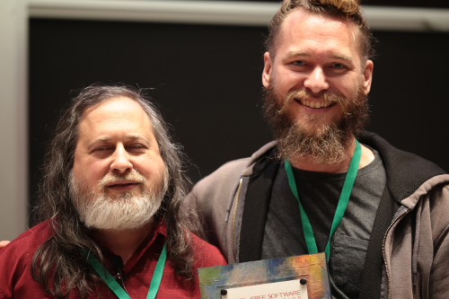 Richard M. Stallman stands to the left of the photo. He has long grey hair and a grey beard. He is wearing a red polo shirt, and a green lanyard with a LibrePlanet conference badge. Conor Schaefer stands to the right. He is taller than Richard. His hair is dirty blond--and tied into a bun. His beard is brown and grey. He is wearing a grey shirt, a grey hoodie, and a green lanyard. He is holding a Free Software Award.