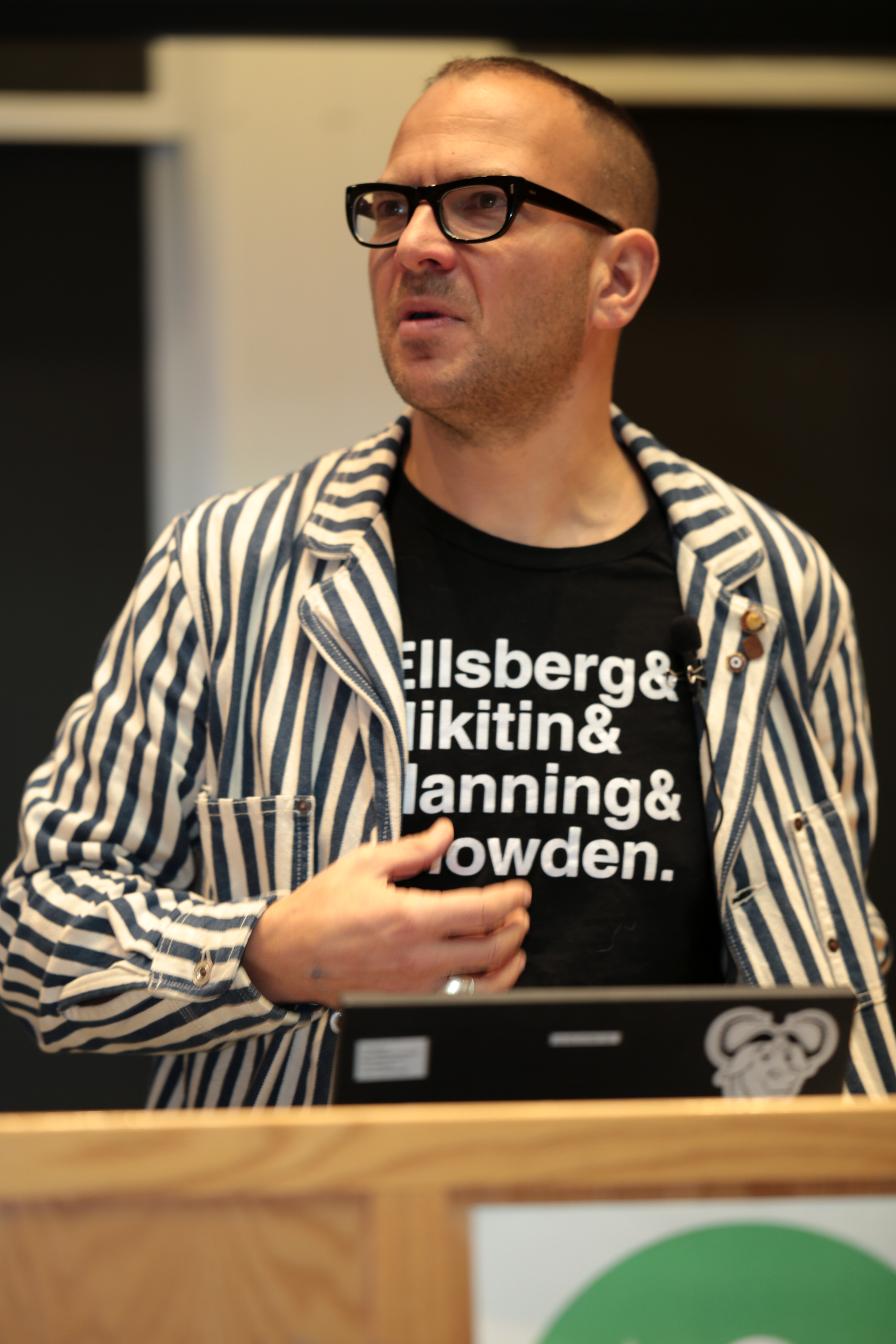 Cory Doctorow stands at a podium in front of a lecture hall. He is wearing a white and navy striped blazer and a black shirt with the text 'Ellsberg & Nikitin & Manning & Snowden.' He has short, dark hair and large, dark glasses.