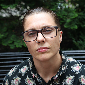 [ A photo of Kade Crockford. They are sitting on a bench in front of some trees, wearing a black shirt with pink and white designs. ]