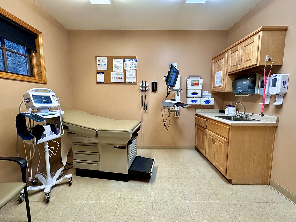 An empty health clinic exam room, complete with exam table, sink, and blood pressure/heart rate machine