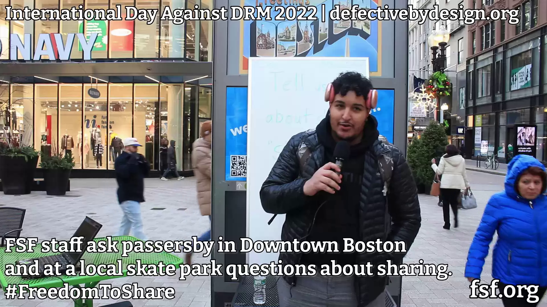 Screenshot from video that says International Day Against DRM 2022 | defectivebydesign.org and has a person with a microphone standing in front of a sign that says Boston. At bottom, it says FSF staff ask passersby in Downtown Boston and at a local skate park questions about sharing. #FreedomToShare