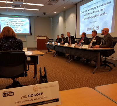 A discussion panel at the event. Six men in suits sit at a long table in front of a projector screen. In the foreground, the author's name plaque is visible on a table.