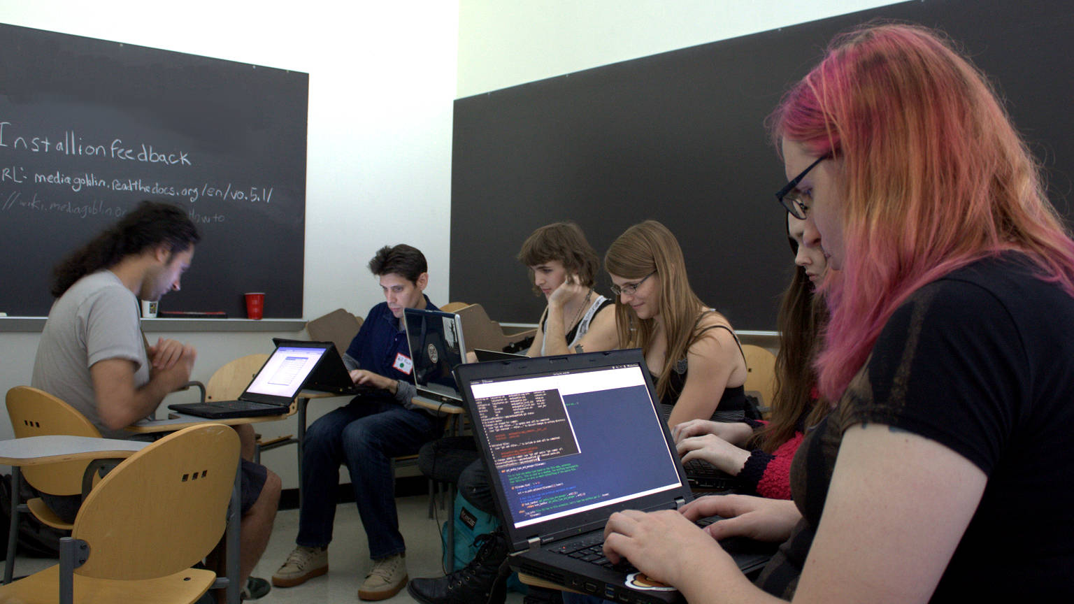 [ A photo of people in a room on laptops. They are sitting at desks, with a clean blackboard in the background on one wall, and on the other written on a bloackboard is the text 