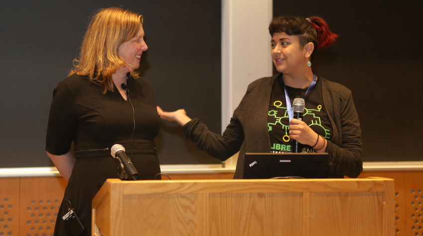 [ Deb Nicholson, wearing a black dress, stands next to Mariah Villarreal, wearing a grey cardigan and LibrePlanet 2018 tshirt. They are both smiling. ]