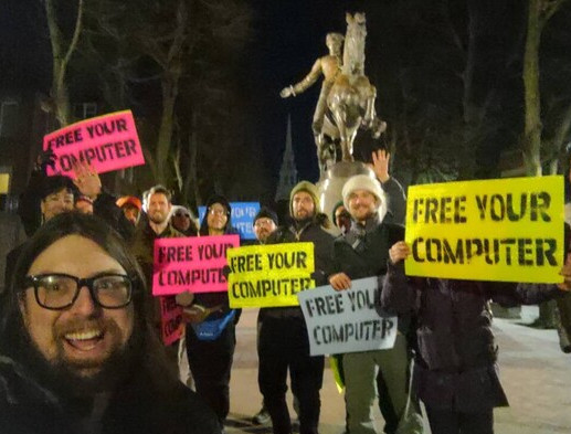 An image of a group of people outside holding signs that say Free your Computer.