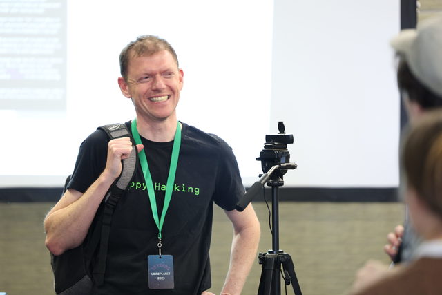 An image of an attendee smiling brightly, wearing a Happy Hacking shirt. He carries a backpack and you can see a projector screen in the background.