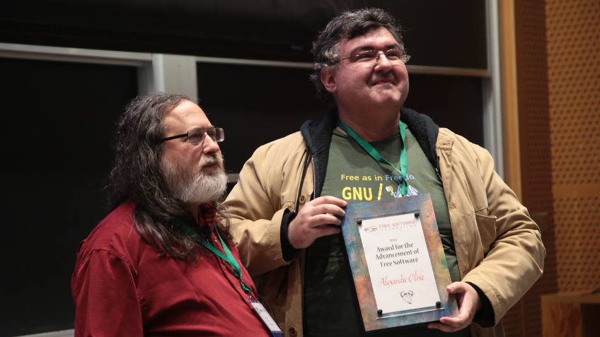 [ A photo of Alexandre Oliva, wearing a green shirt and a tan jacket, with Richard Stallman, wearing a red shirt. Oliva is holding a plaque that reads 