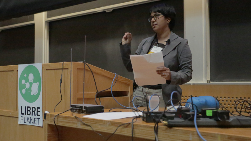 [ Sumana Harihareswara speaking at a podium at LibrePlanet 2016. Between her and the camera is a table strewn with cables and computer equipment. ]