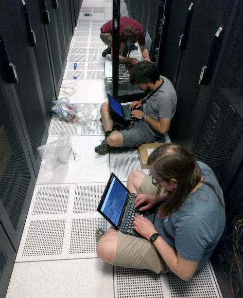 Members of the FSF tech team, three men with laptops, sitting on the floor in a hallway, working on migration to a new colocation facility in June 2019.